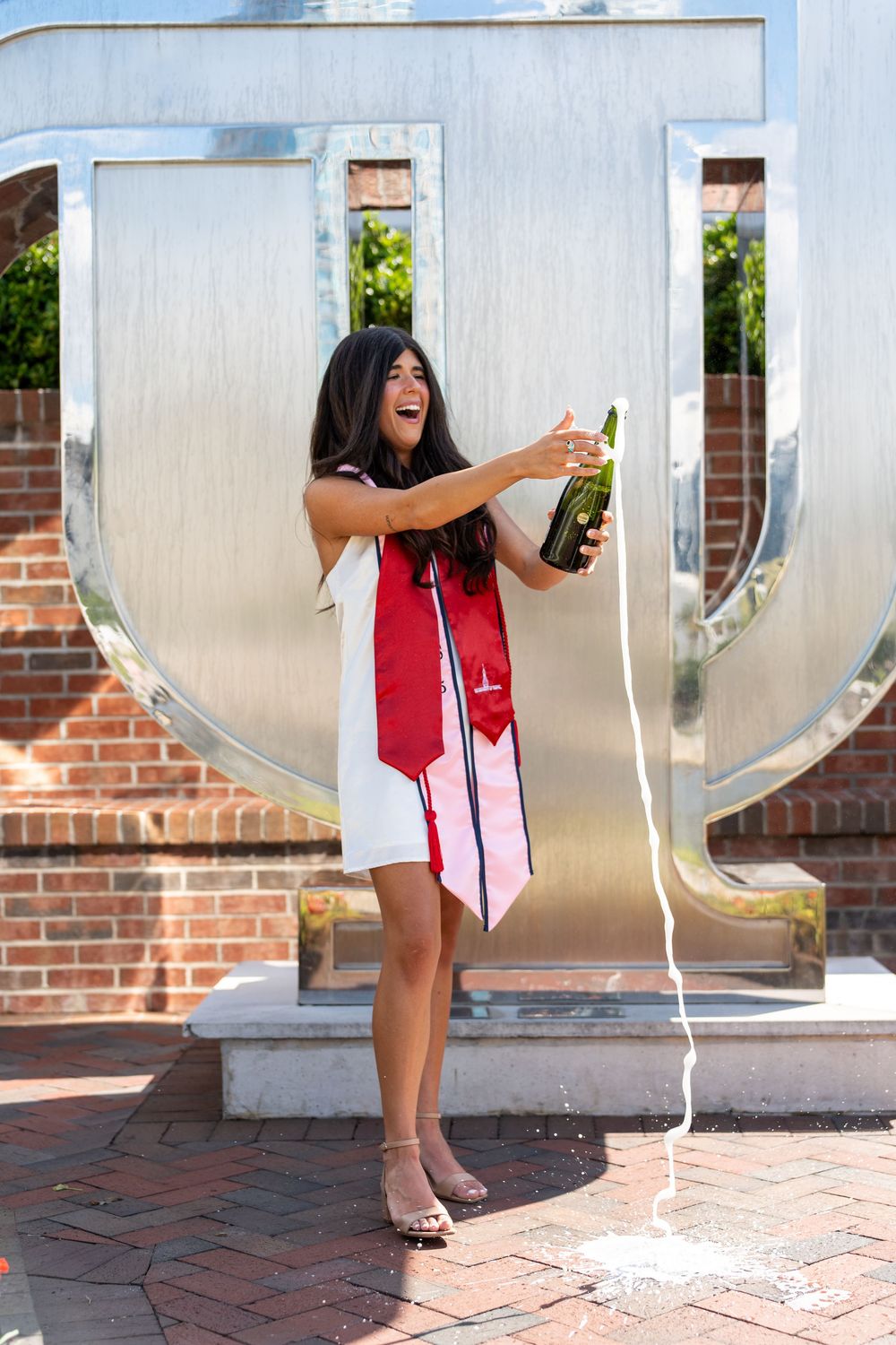 A graduate in red and white regalia pops open a champagne bottle while celebrating in front of a large U-shaped monument.