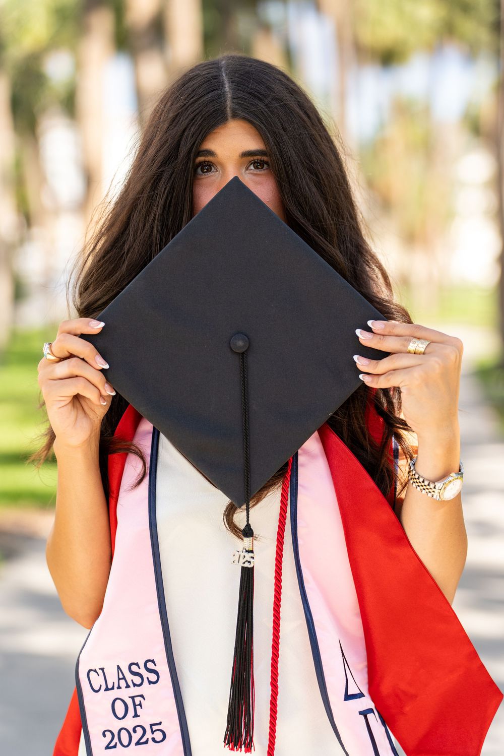 Graduate holding black mortarboard with Class of 2025 stole outdoors with palm trees in background.