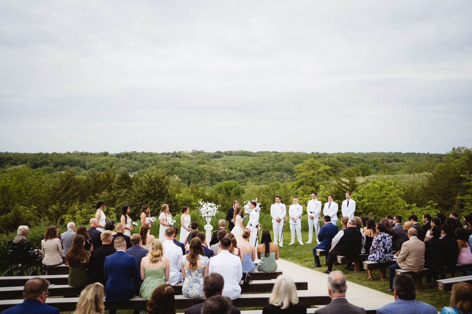 Wedding ceremony taking place outdoors with guests seated on wooden benches overlooking a green countryside landscape.