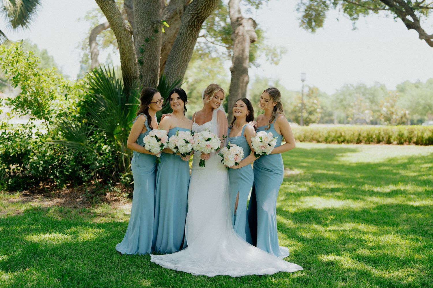 A bridal party in flowing light blue bridesmaid dresses pose on a sunny lawn with palm trees and greenery.