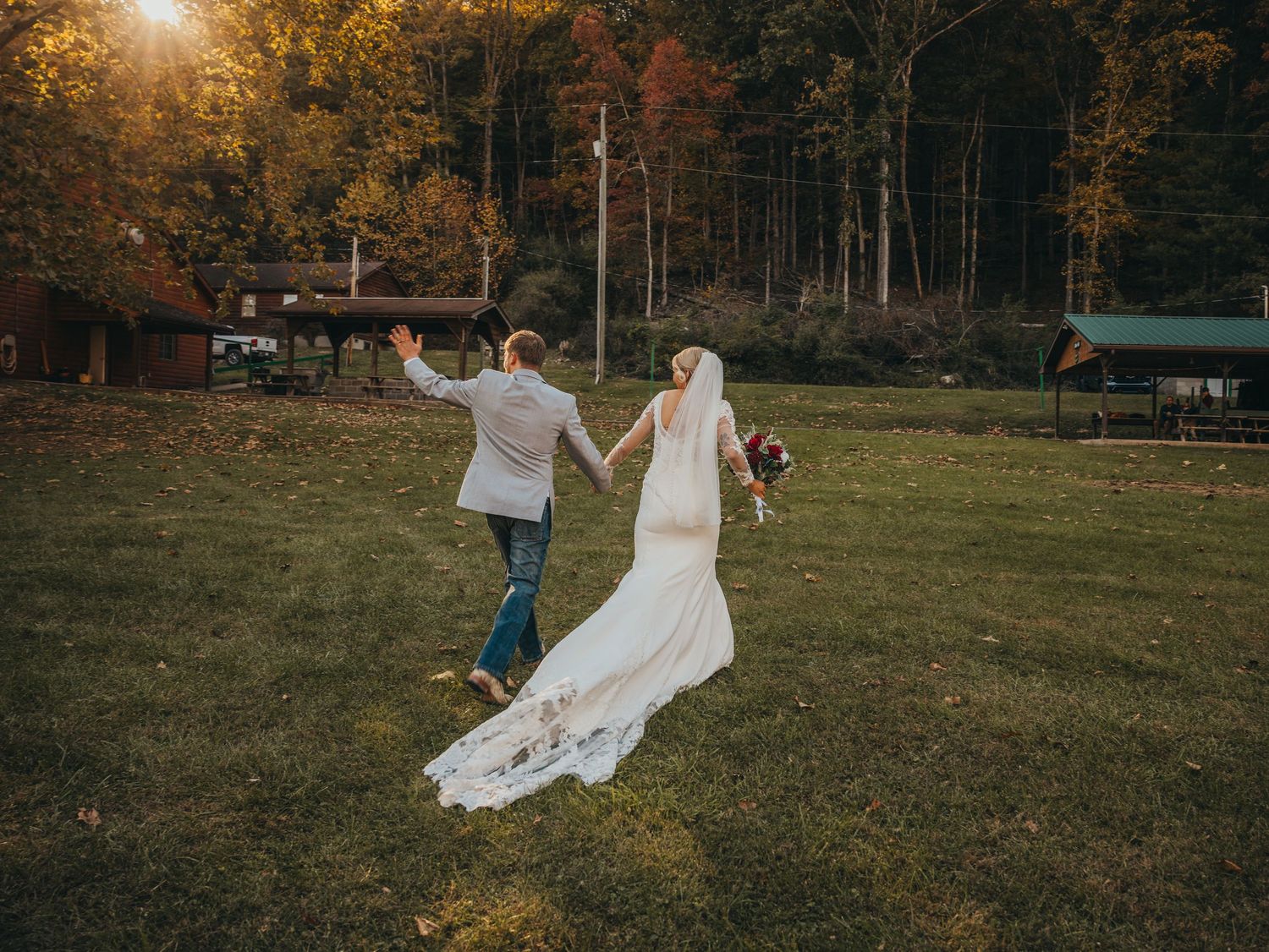 Newlyweds walk hand in hand across field at sunset with trailing wedding dress.