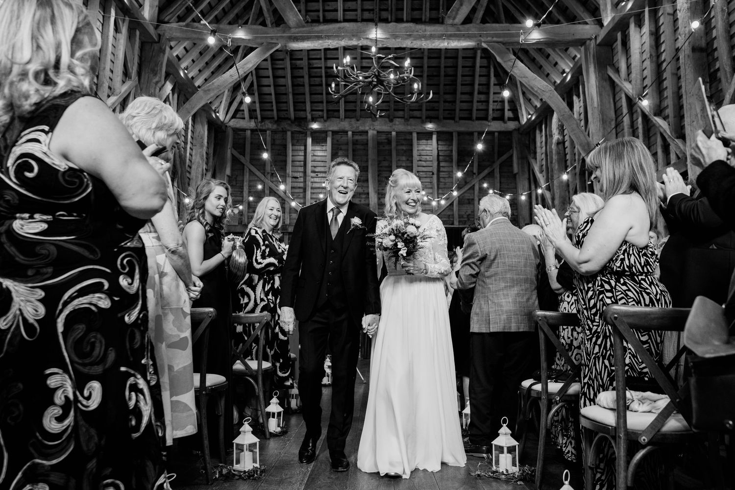 A black and white photograph of a wedding celebration inside a rustic barn decorated with lanterns and twinkling lights.