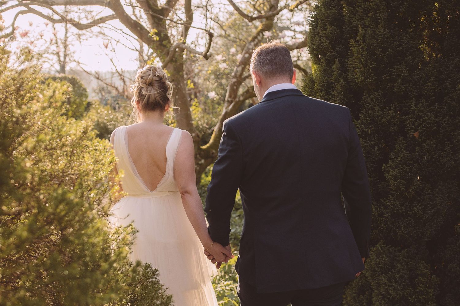 The bride and groom walk together on a sunlit pathway surrounded by lush greenery.