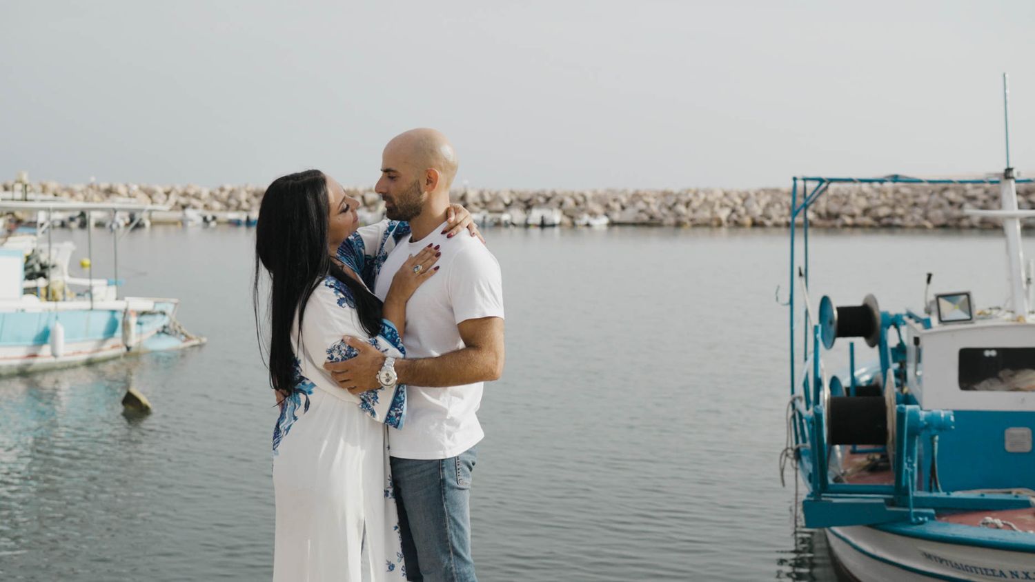 A couple stands close together on a dock with boats and calm water in the background at a marina.