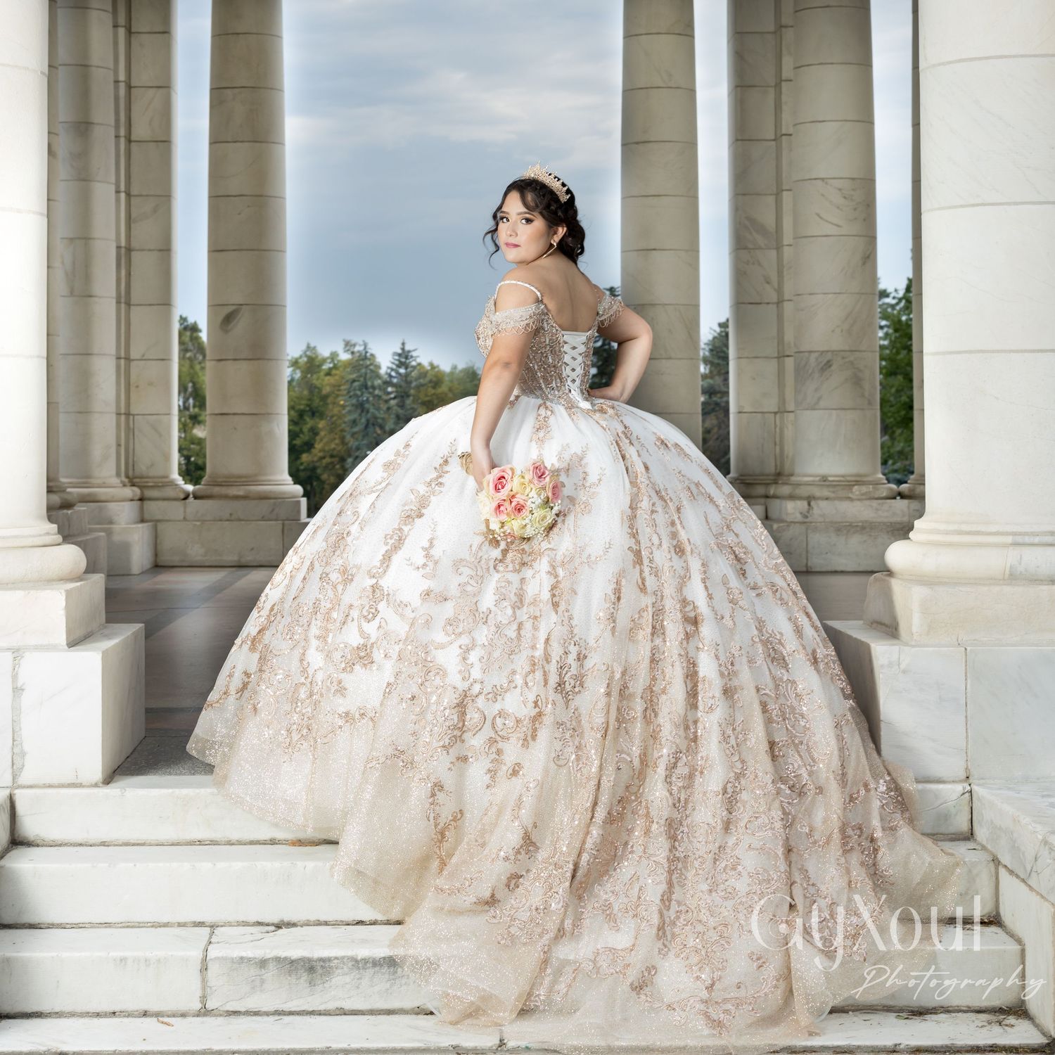 A bride in a glamorous lace ball gown poses on marble steps between classic stone columns holding a peach bouquet.