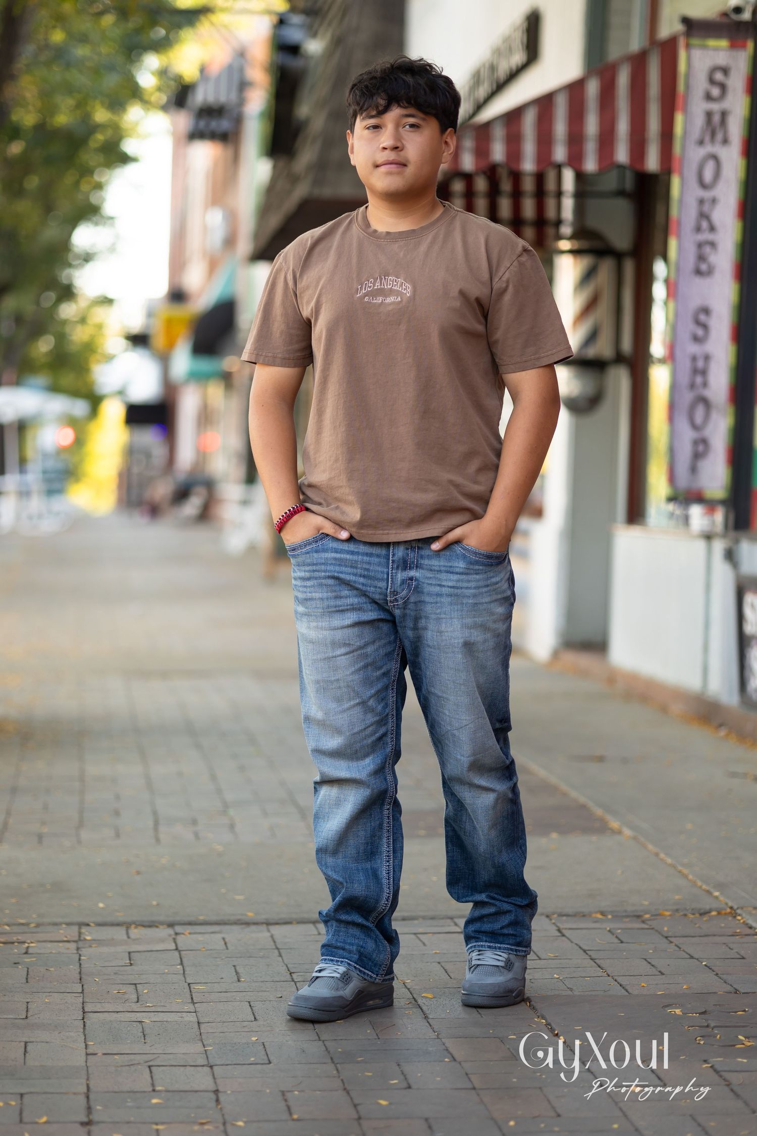 Person in casual brown t-shirt and blue jeans standing on brick sidewalk in front of downtown storefronts.