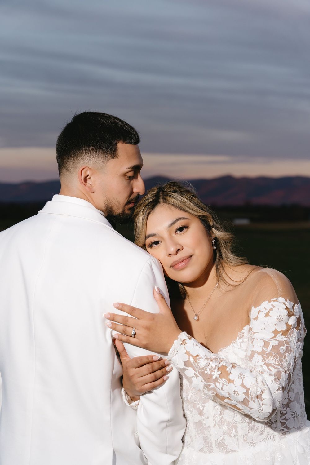 Romantic sunset portrait of couple embracing in formal wedding attire against mountain backdrop.