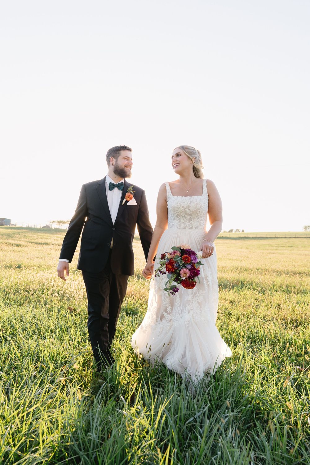 Newlywed couple walks through golden wheat field at sunset in formal wedding attire with red bouquet.