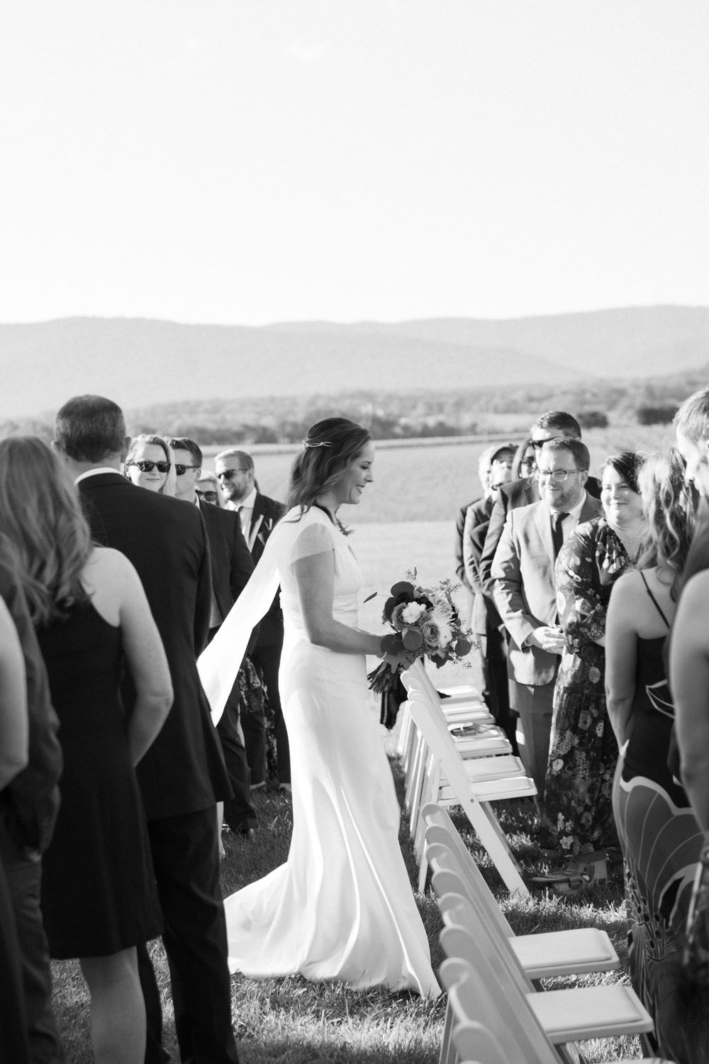 A wedding ceremony processional with guests seated in rows on an outdoor hillside.