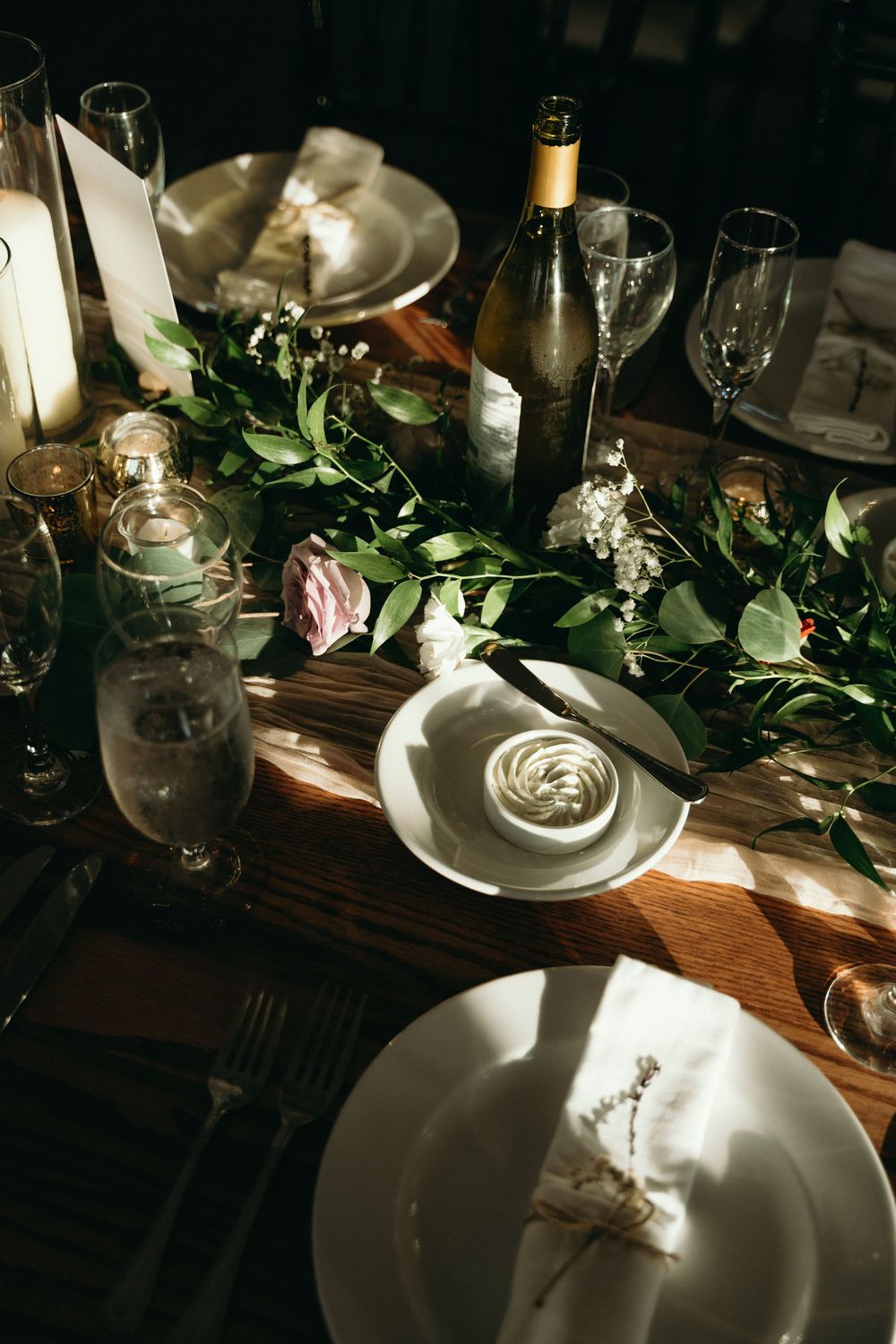 Elegant wedding table setting with greenery, candles, and wine bottles in dramatic lighting.