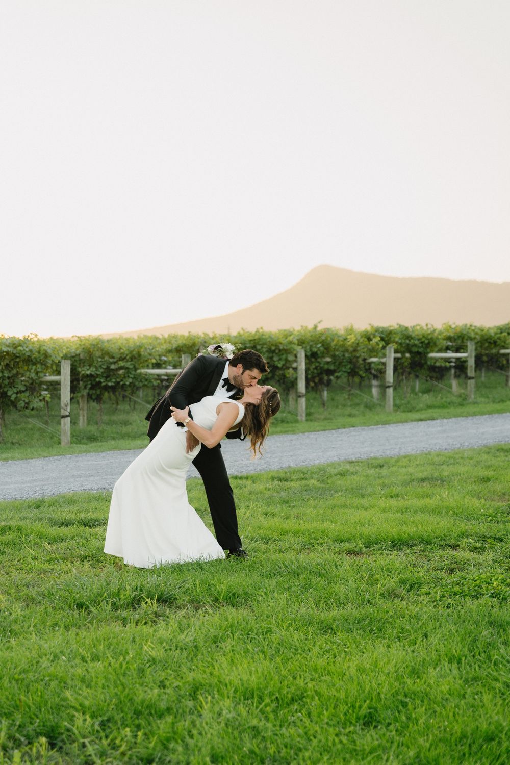 A couple shares a romantic dip kiss on a grassy path with rolling hills in the background at a vineyard.