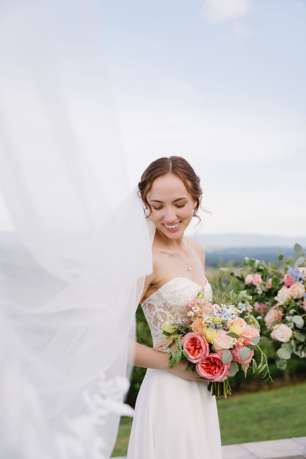Bride in a strapless white gown smiles while holding a coral and peach floral bouquet against a scenic mountain backdrop.