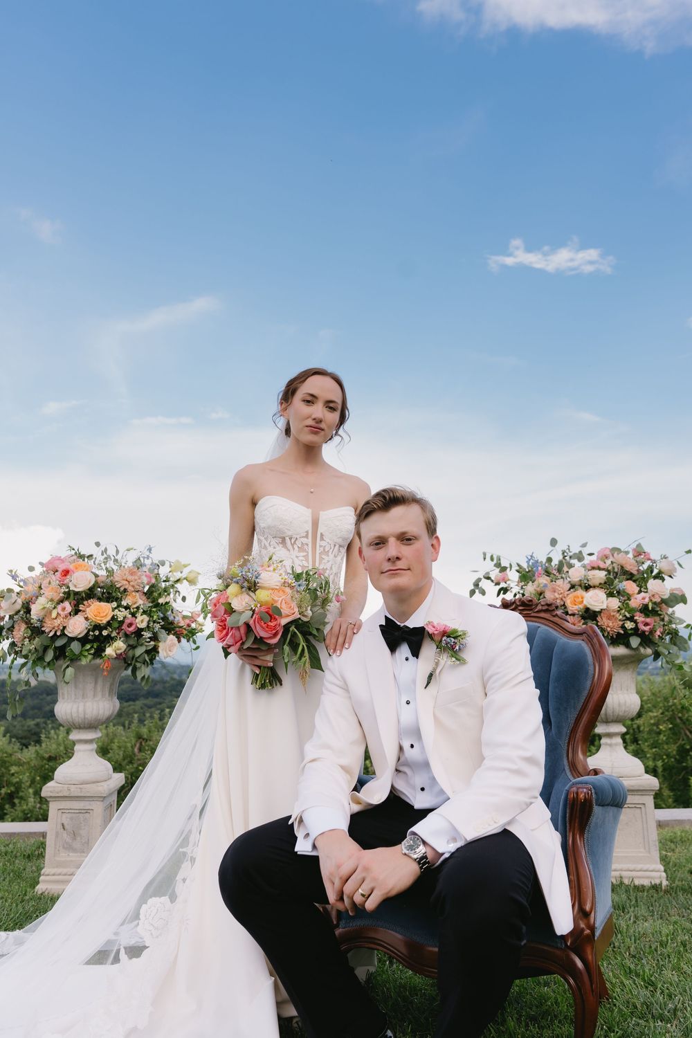 Elegant wedding portrait with couple posed by stone urns filled with coral flowers on scenic hilltop terrace.