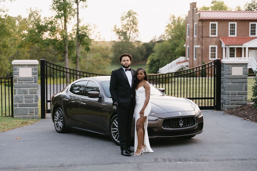 A couple poses in formal attire next to a black Maserati luxury car in front of an elegant gated driveway.
