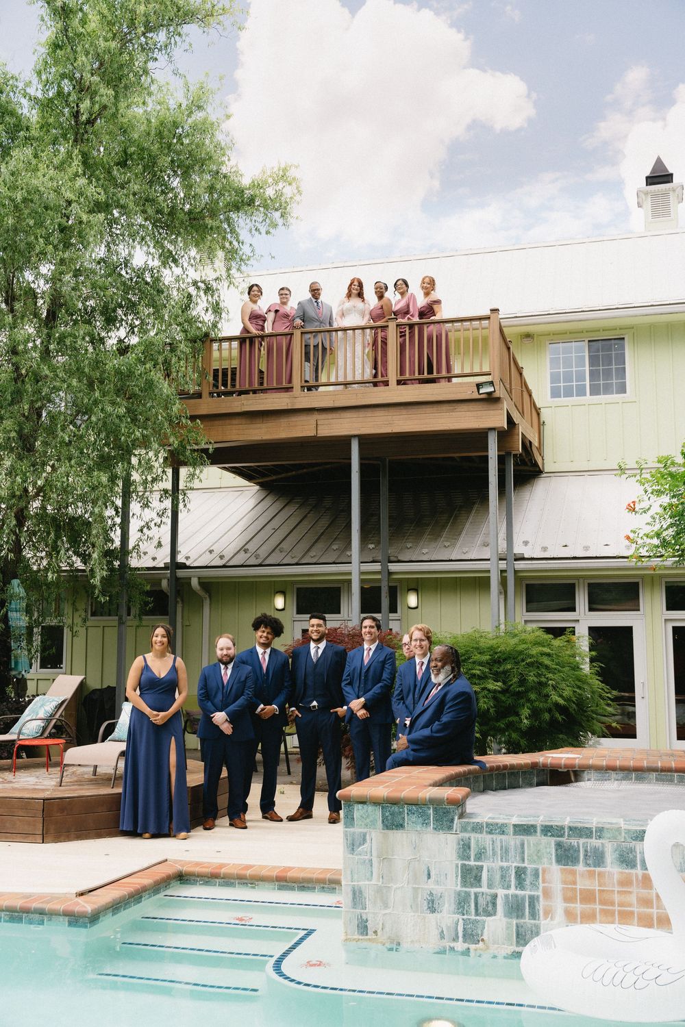 Wedding party stands on a modern wooden deck overlooking a swimming pool during an outdoor celebration.