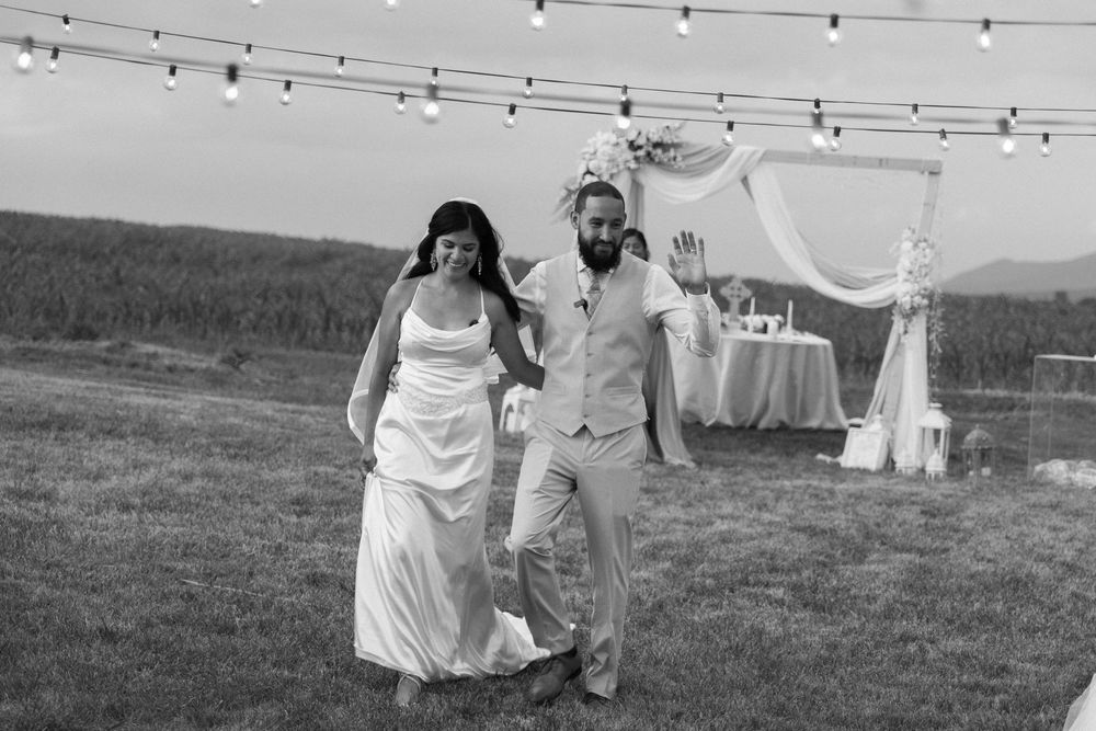 Newlyweds walking joyfully under string lights at outdoor wedding reception in black and white.