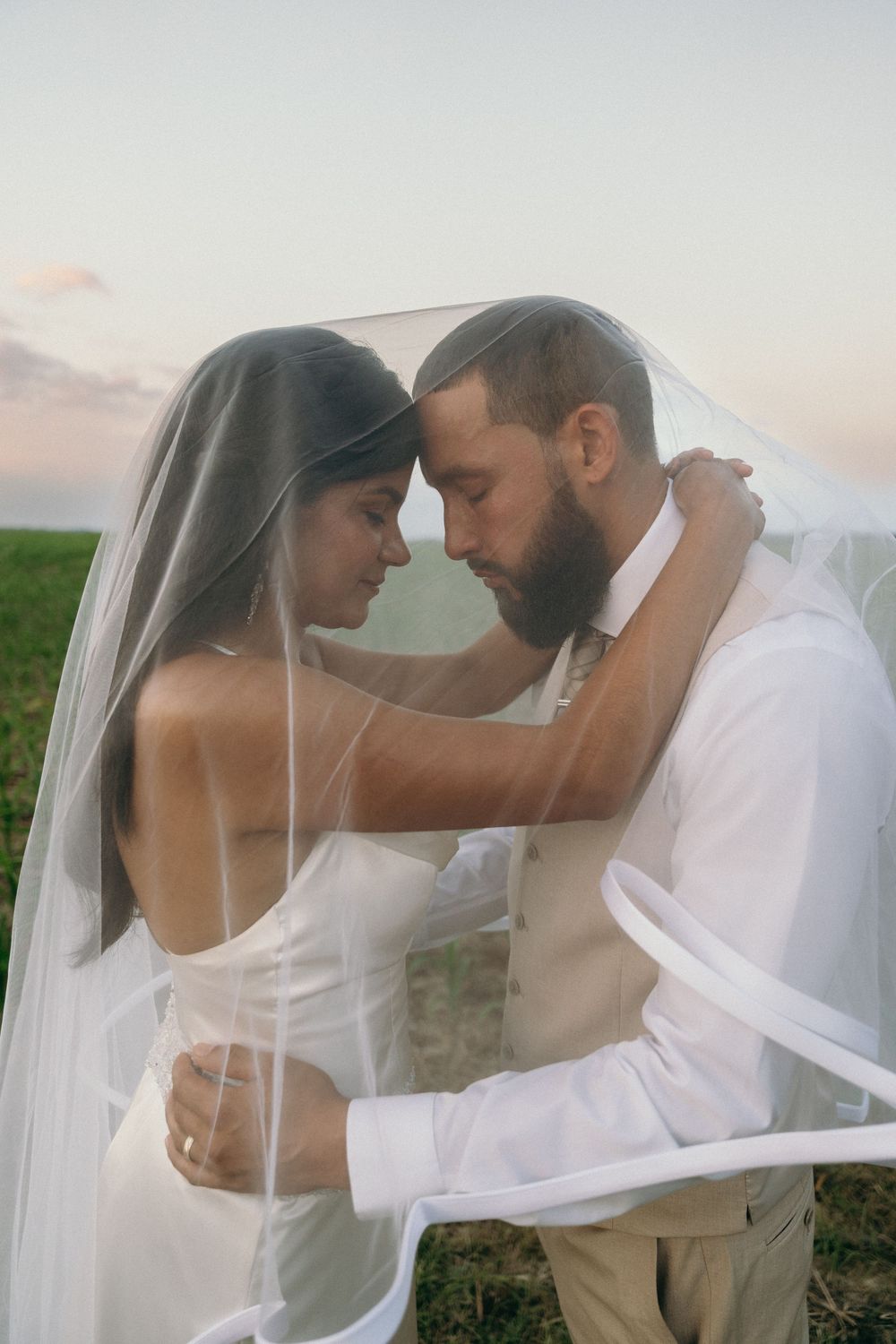 Intimate outdoor wedding portrait of a bride and groom sharing a tender moment under a flowing veil at sunset.