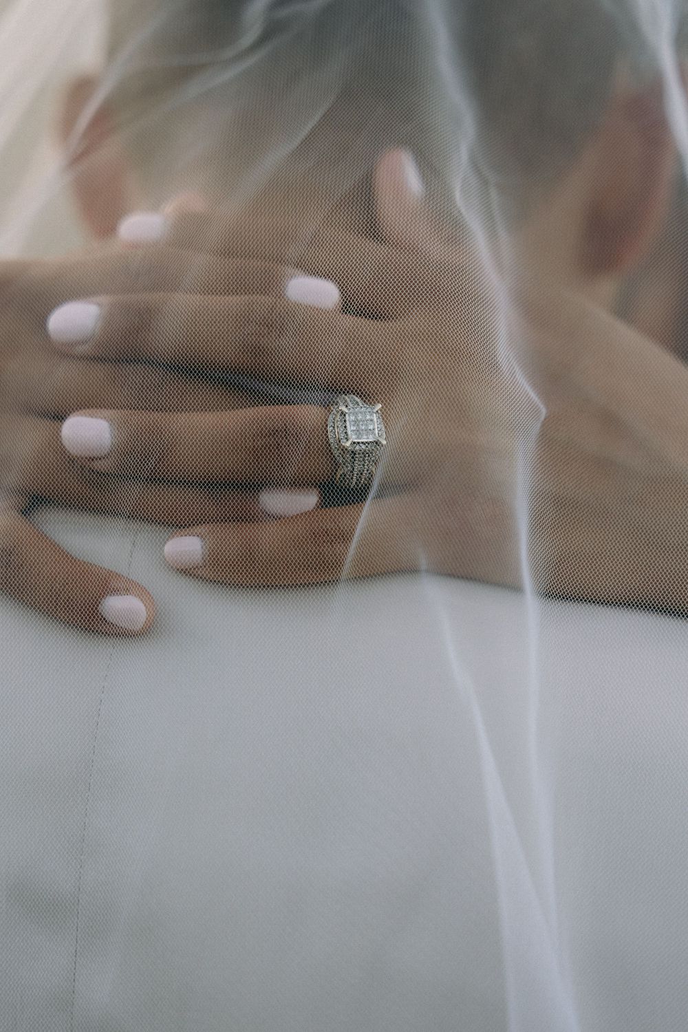 Close-up detail shot of hands with white manicure and sparkling diamond engagement ring under wedding veil.