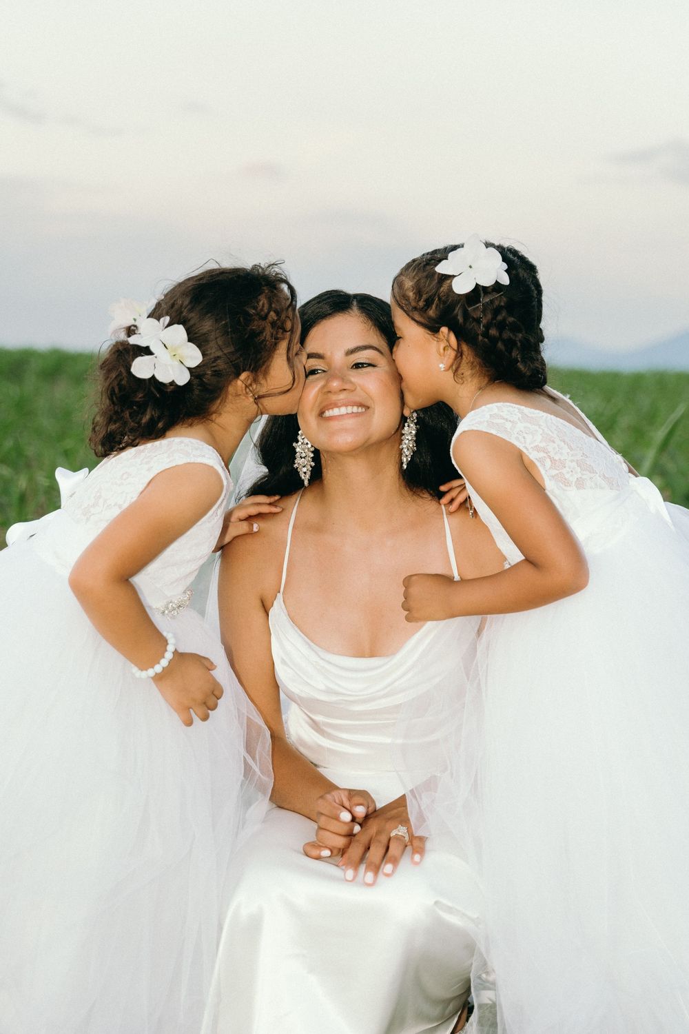 Sweet moment captured as two flower girls in white dresses kiss the bride on her cheeks.