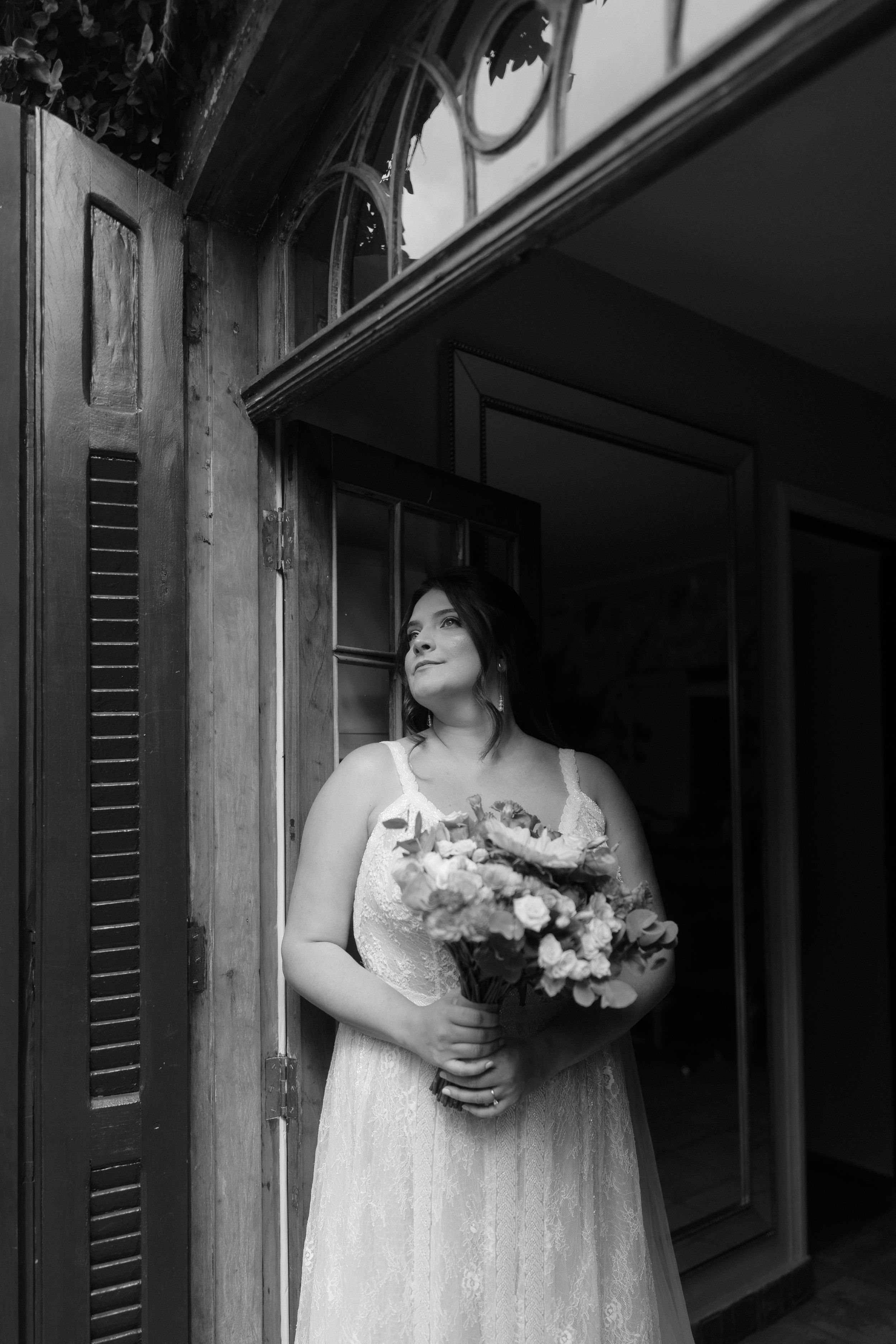 Black and white portrait of person in wedding dress holding bouquet by vintage window.