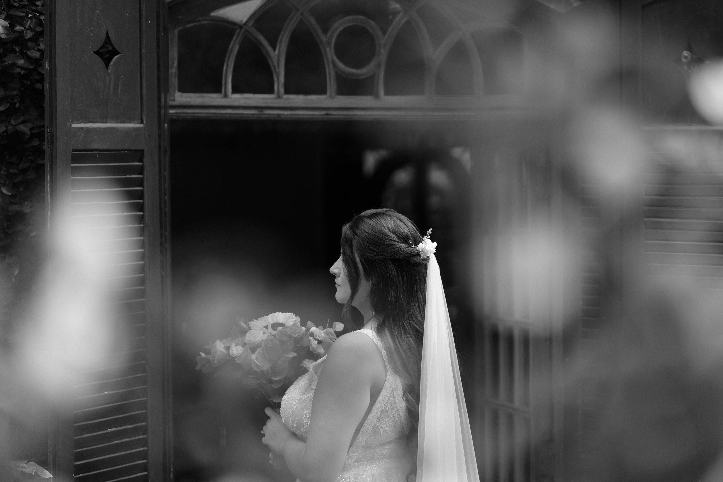 Artistic black and white photo of bride with bouquet through window reflection.