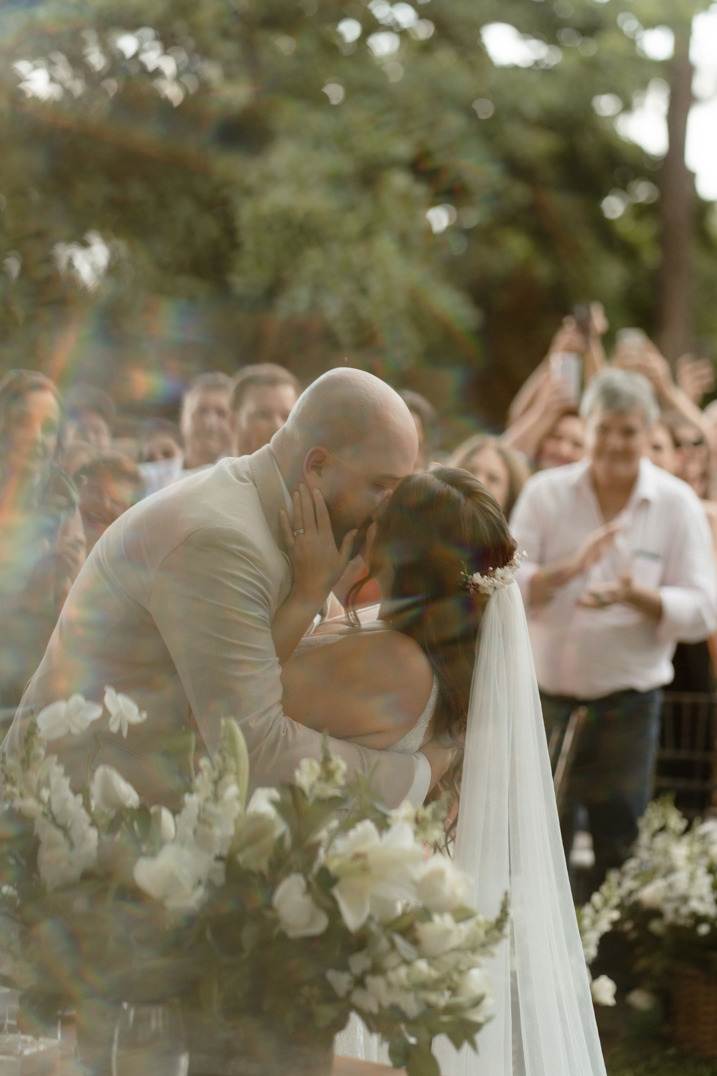 Romantic wedding kiss surrounded by white floral arrangements and cheering guests.