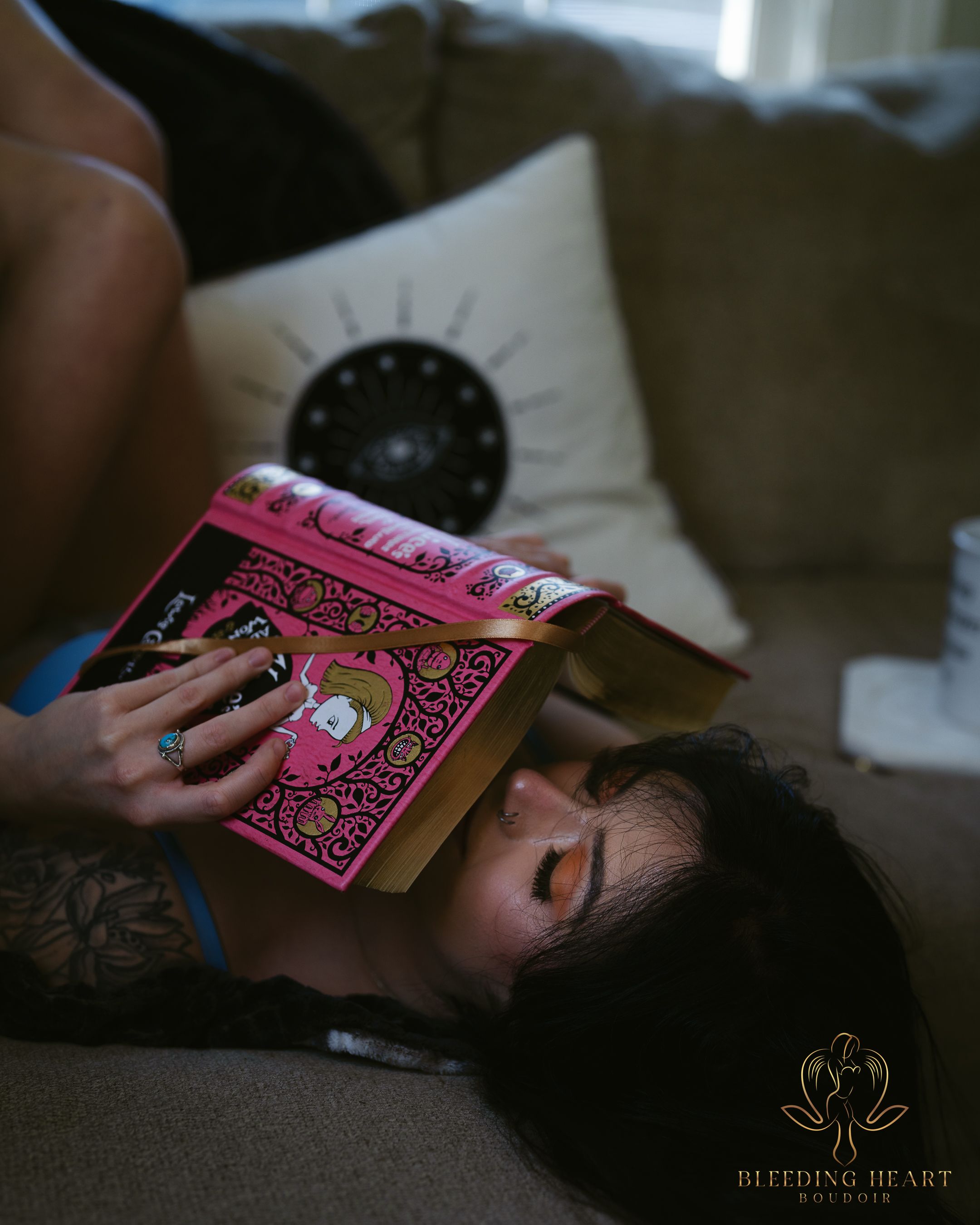 Someone lies on a brown couch reading a pink hardcover book, with a decorative pillow visible in the background.