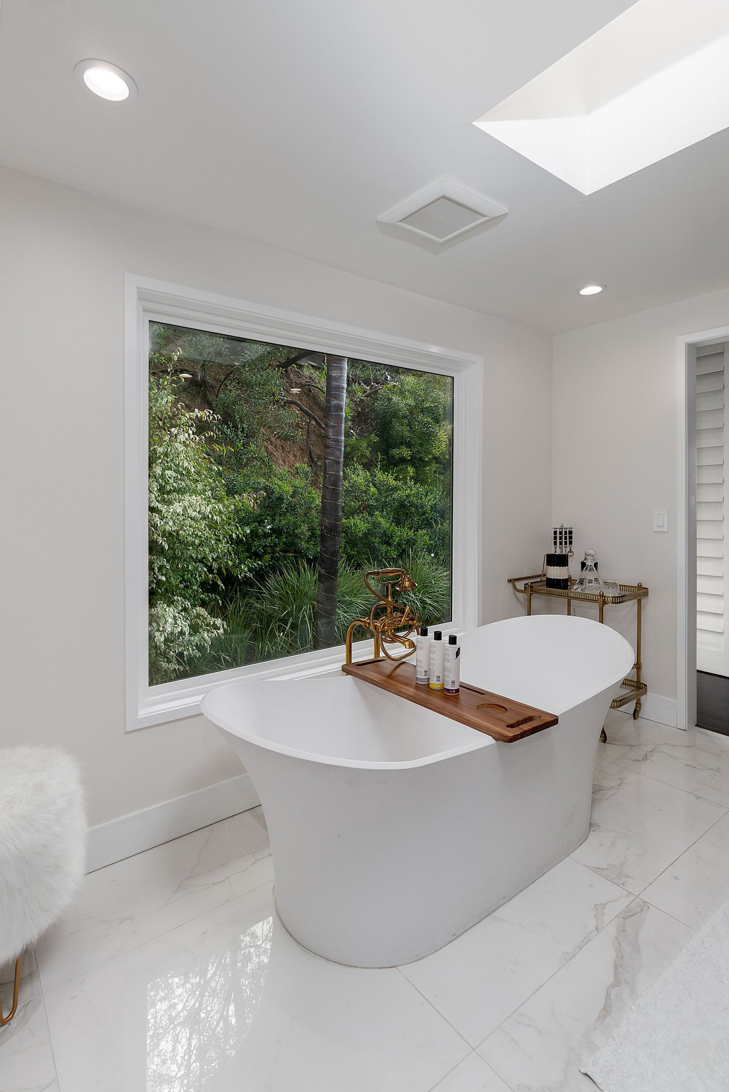 Modern bathroom with freestanding white tub, wooden shelf, and large window overlooking lush greenery.