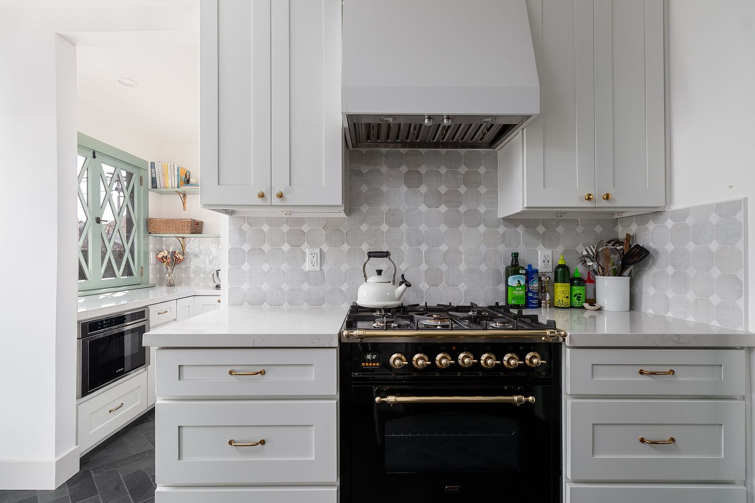 Modern white kitchen with black stove and brass hardware features geometric tile backsplash and marble countertops.