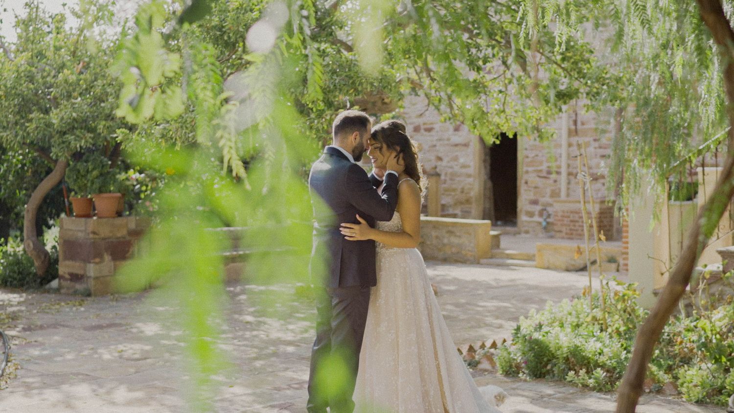A romantic couple embraces in a garden courtyard with stone walls and green foliage framing the scene.