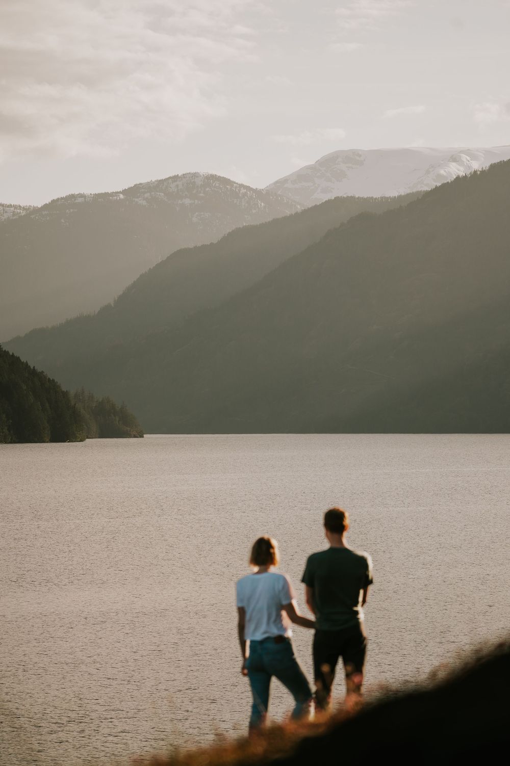 Engaged couple looking out at views of Comox Lake Bluffs during golden hour holding hands with lake and mountain views in the background