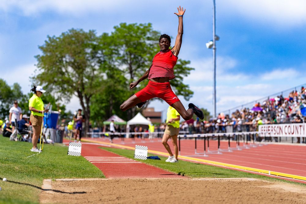 Athlete performs long jump at outdoor track meet, soaring through air in red uniform on sunny day.