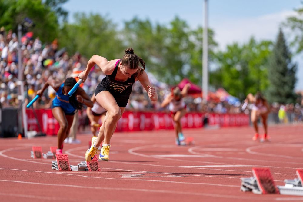 Female sprinter exploding off starting blocks on red track at outdoor athletics competition with crowd in background.