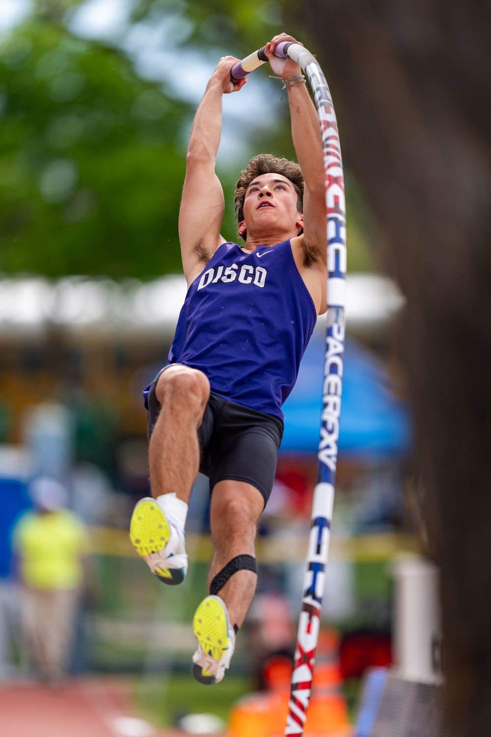 Male athlete in blue OJSCO jersey performs pole vault at outdoor track meet, gripping pole mid-jump.