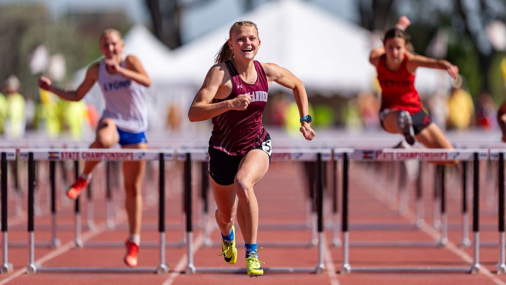 Female athlete smiling while clearing a hurdle during a track and field championship race, competitors visible behind her.