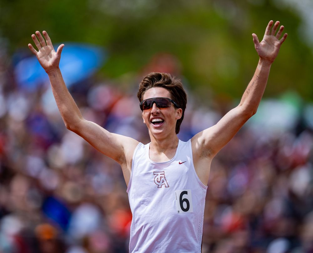 Athlete in white jersey and sunglasses celebrates with arms raised in victory during outdoor competition.
