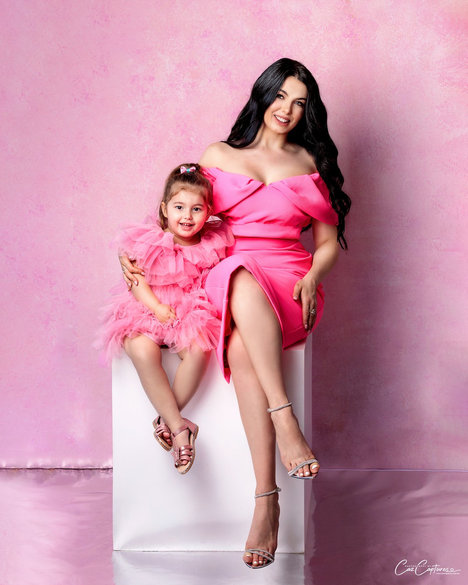 Mother and daughter smiling in pink themed set sitting on white posing block.