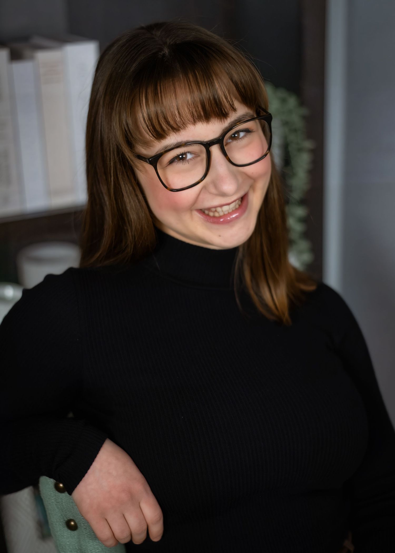 Professional headshot of a person in a black off-shoulder top with long brown hair smiling warmly against a neutral background.