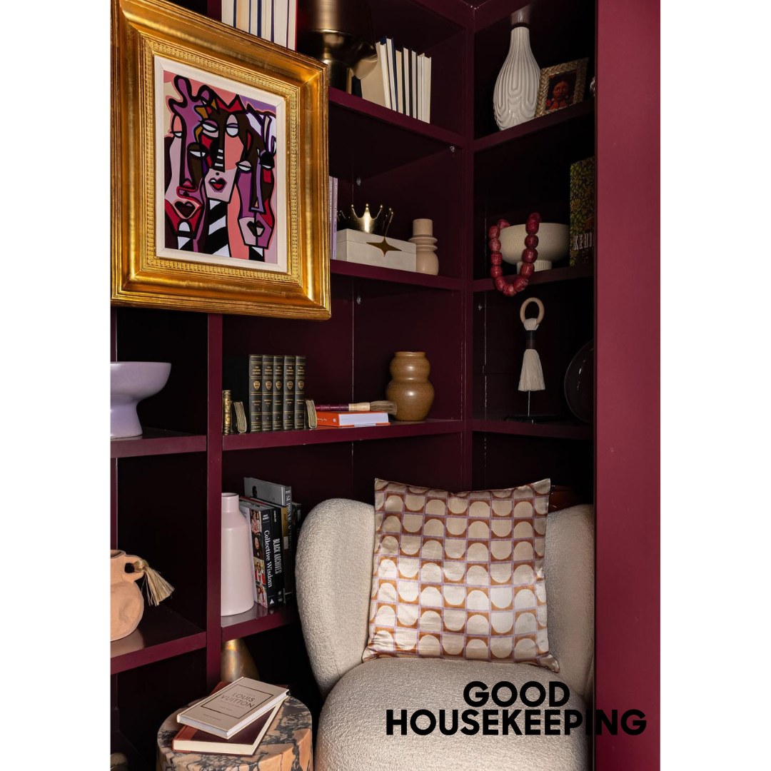 Heather Ison Photography for Good Housekeeping. A cozy reading nook with burgundy built-in shelves, a comfortable armchair, framed artwork.