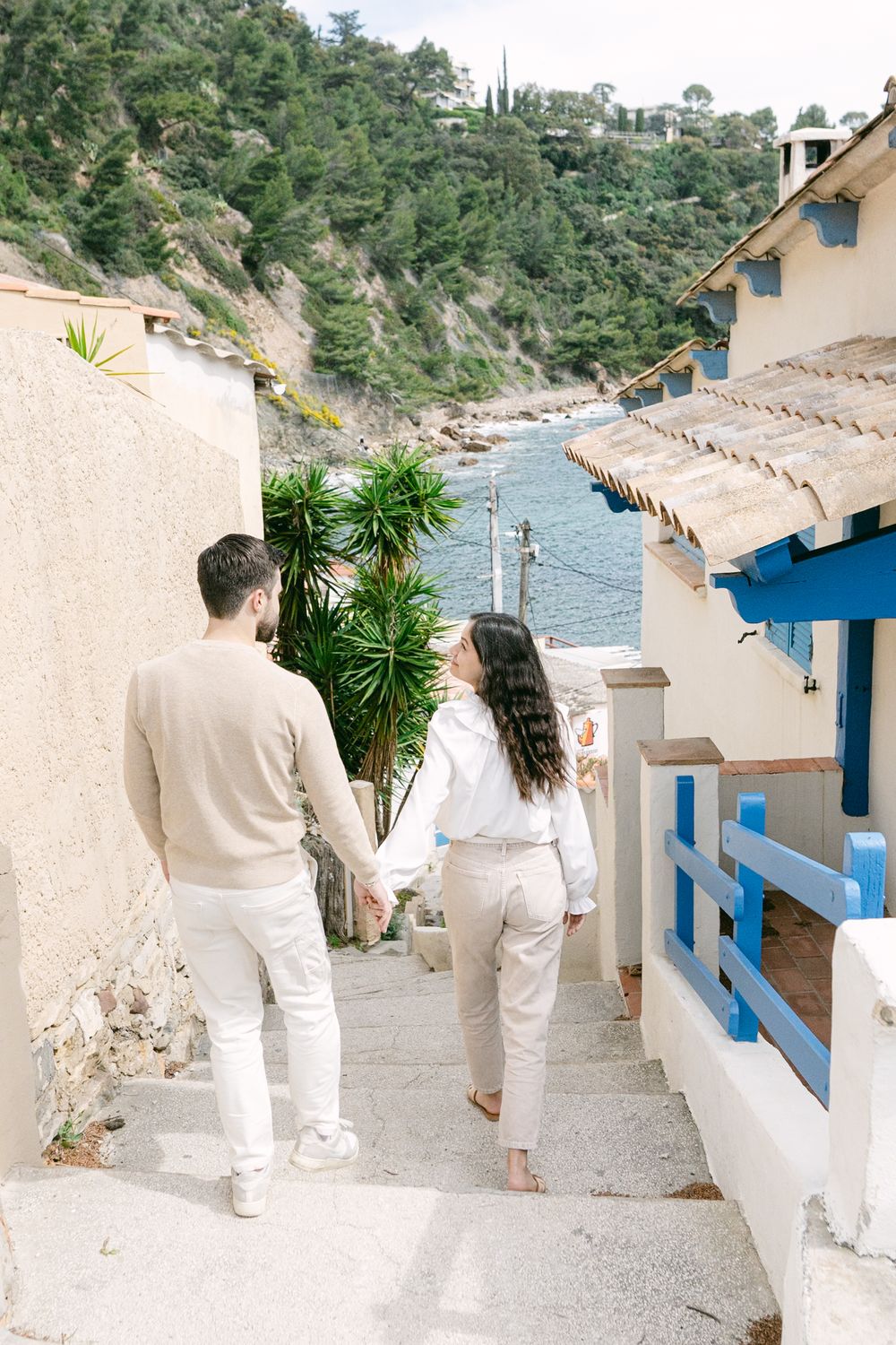 A couple in matching white outfits walks down Mediterranean steps with ocean views and blue accents.