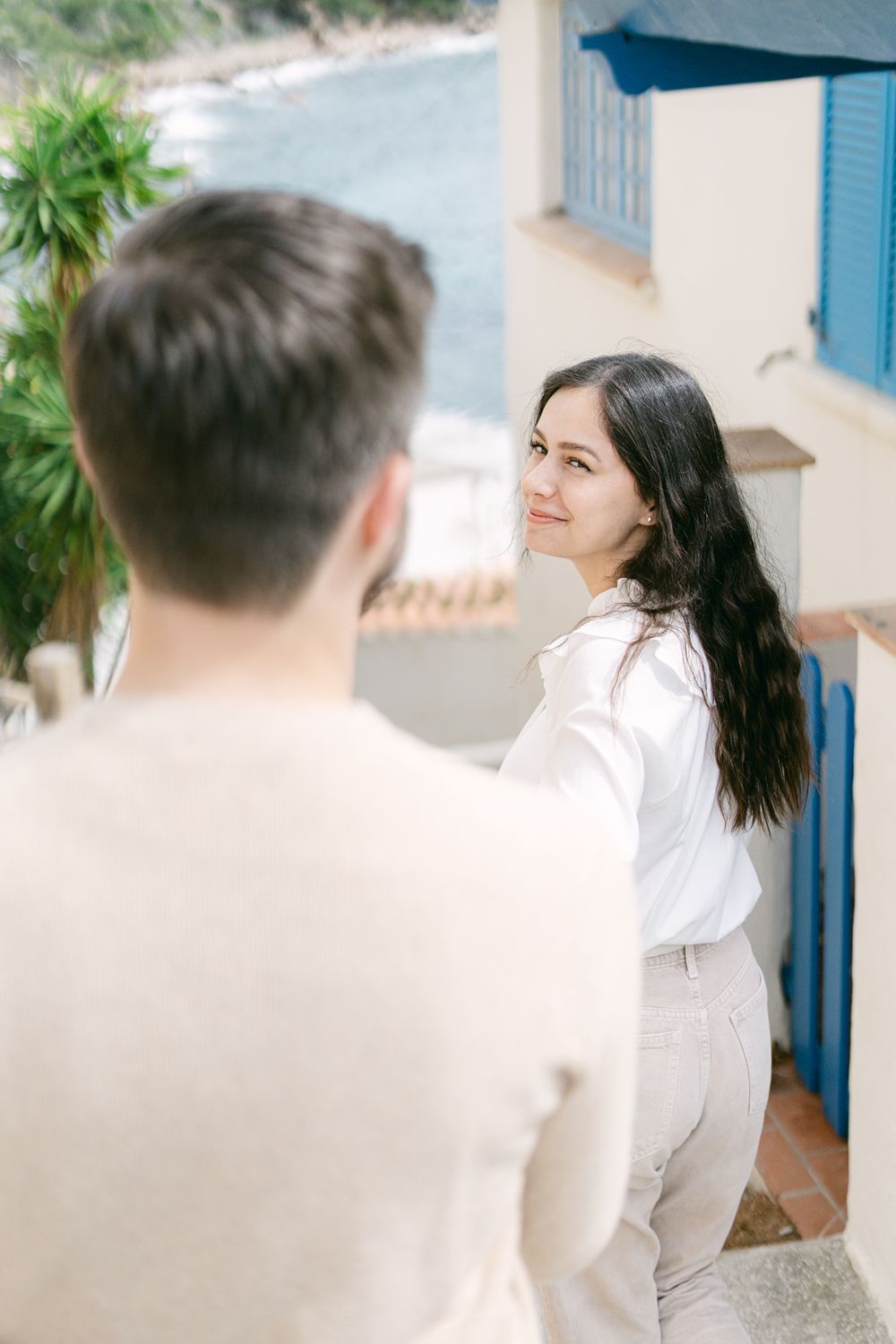Two people in white outfits share a playful moment on an outdoor patio with blue and white architectural details.