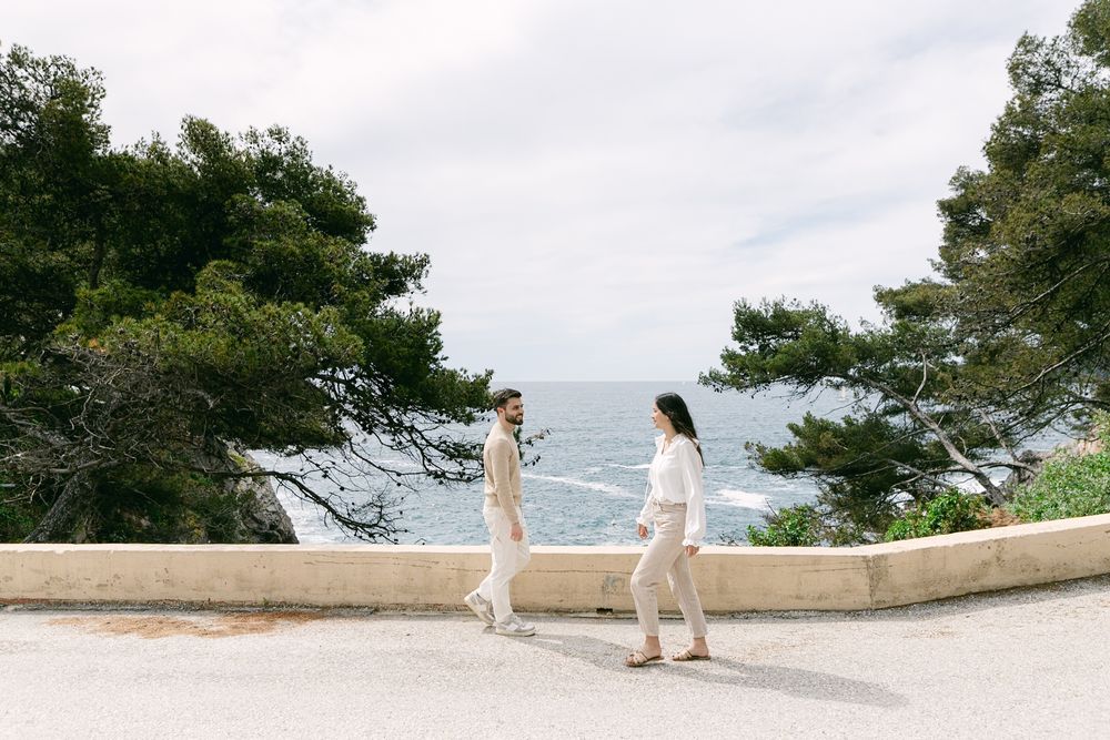 Two people in white outfits walk along a coastal path with evergreen trees and ocean views in the background.