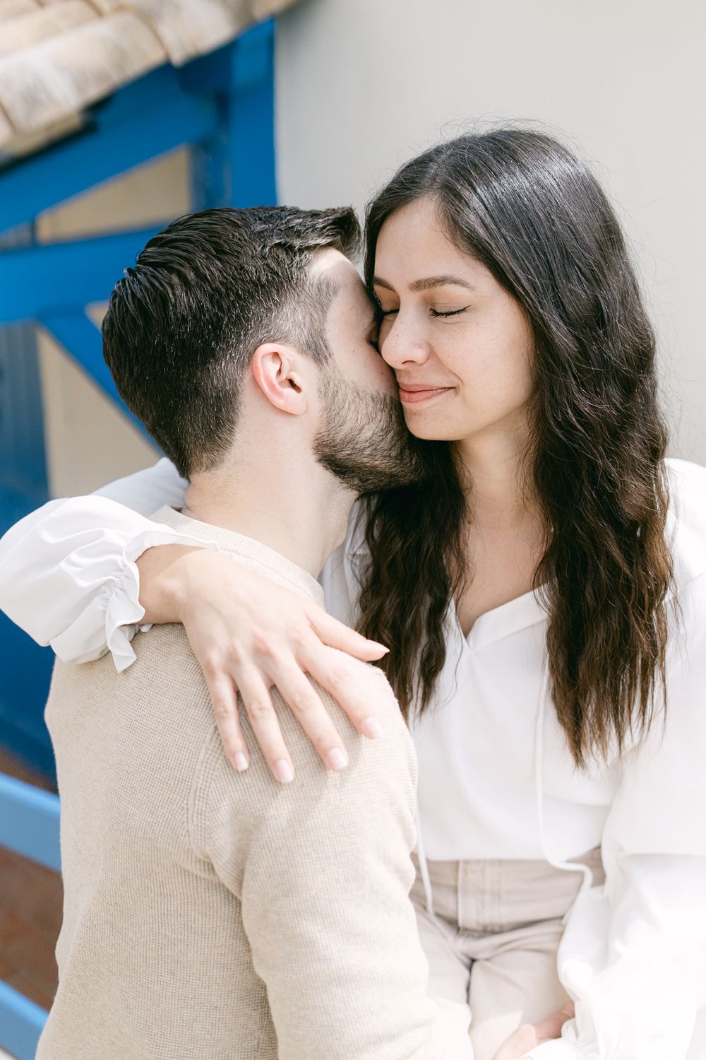 A tender romantic moment shows a couple in white and beige clothing embracing intimately against a light background.