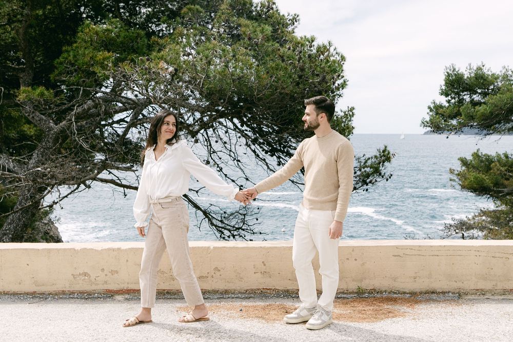 Two people in light colored outfits hold hands while walking along a coastal path with ocean and trees in background.