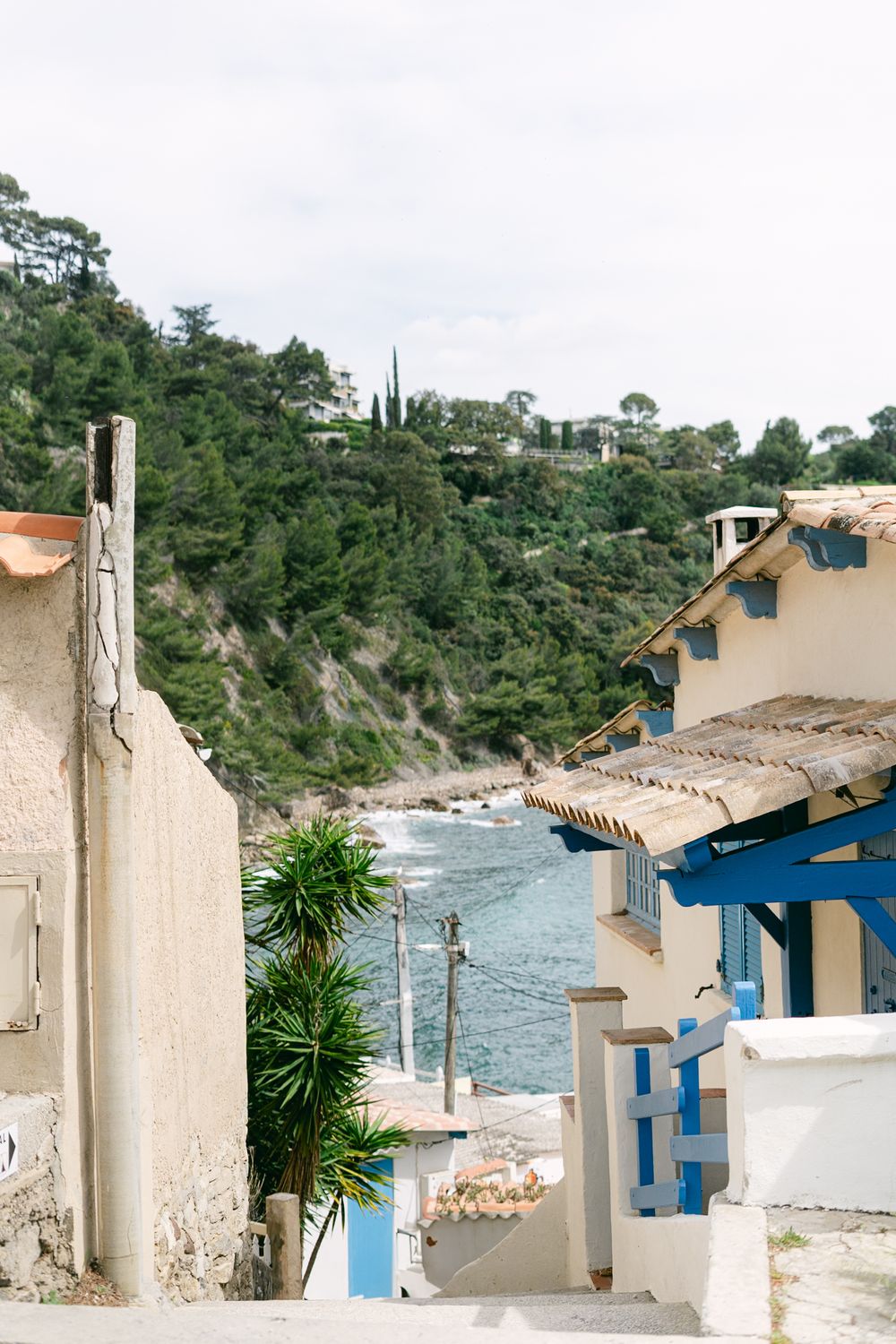 A charming Mediterranean coastal street with white buildings, blue accents, and a view of the rocky coastline and sea.