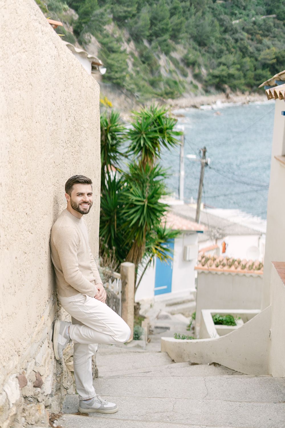 A scenic coastal hillside path with lush palm plants and white buildings overlooking a Mediterranean beach.