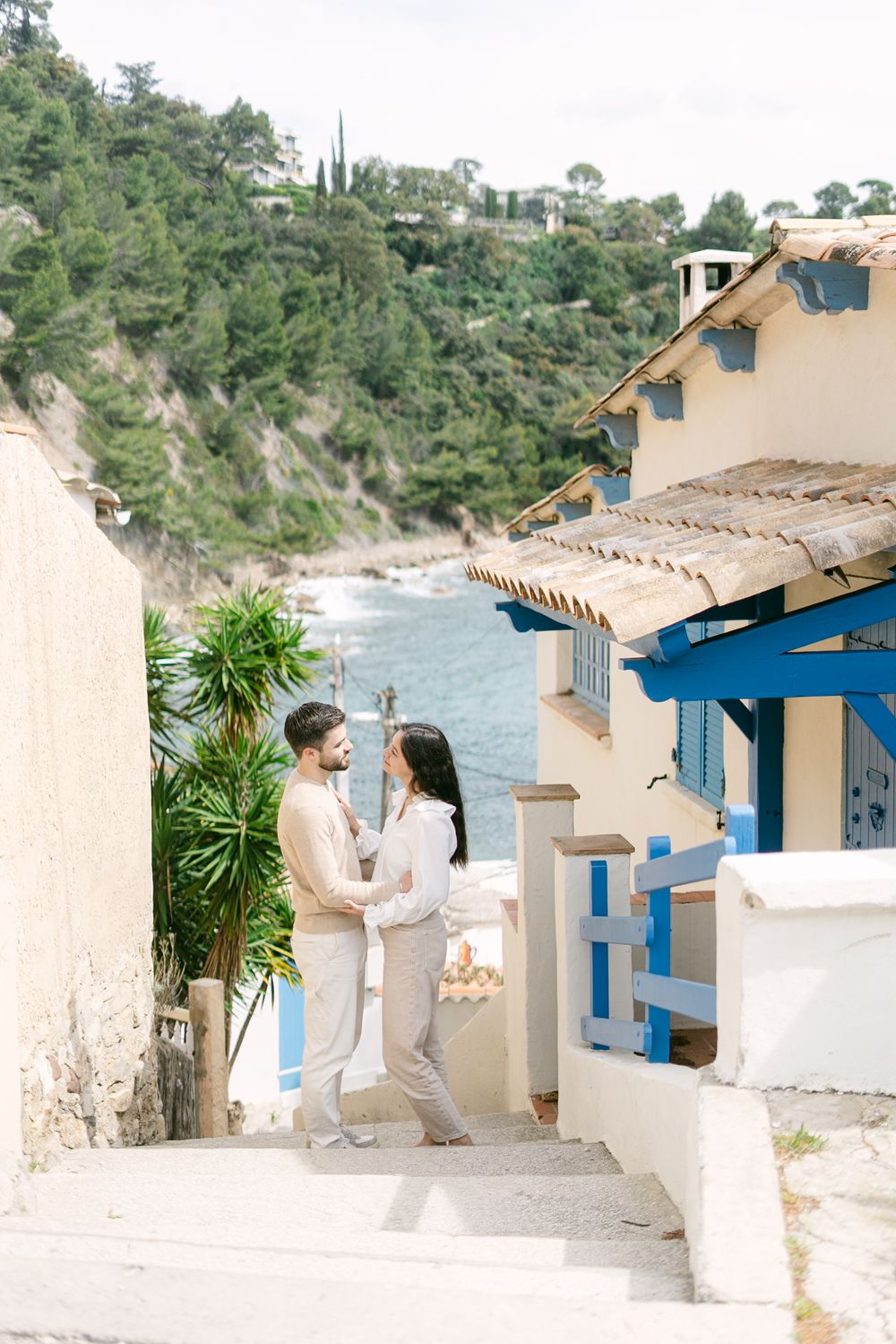 A couple stands on white Mediterranean steps overlooking a coastal cove with blue trim buildings and water views.