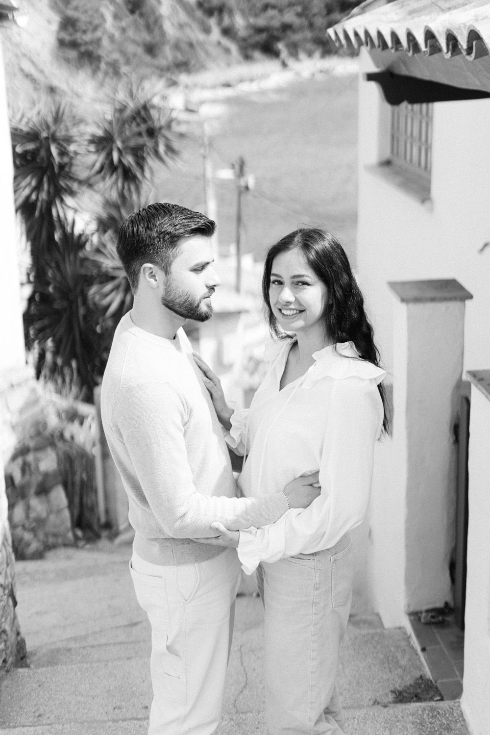 Black and white portrait of couple in coordinated white outfits posing outside Spanish-style building.