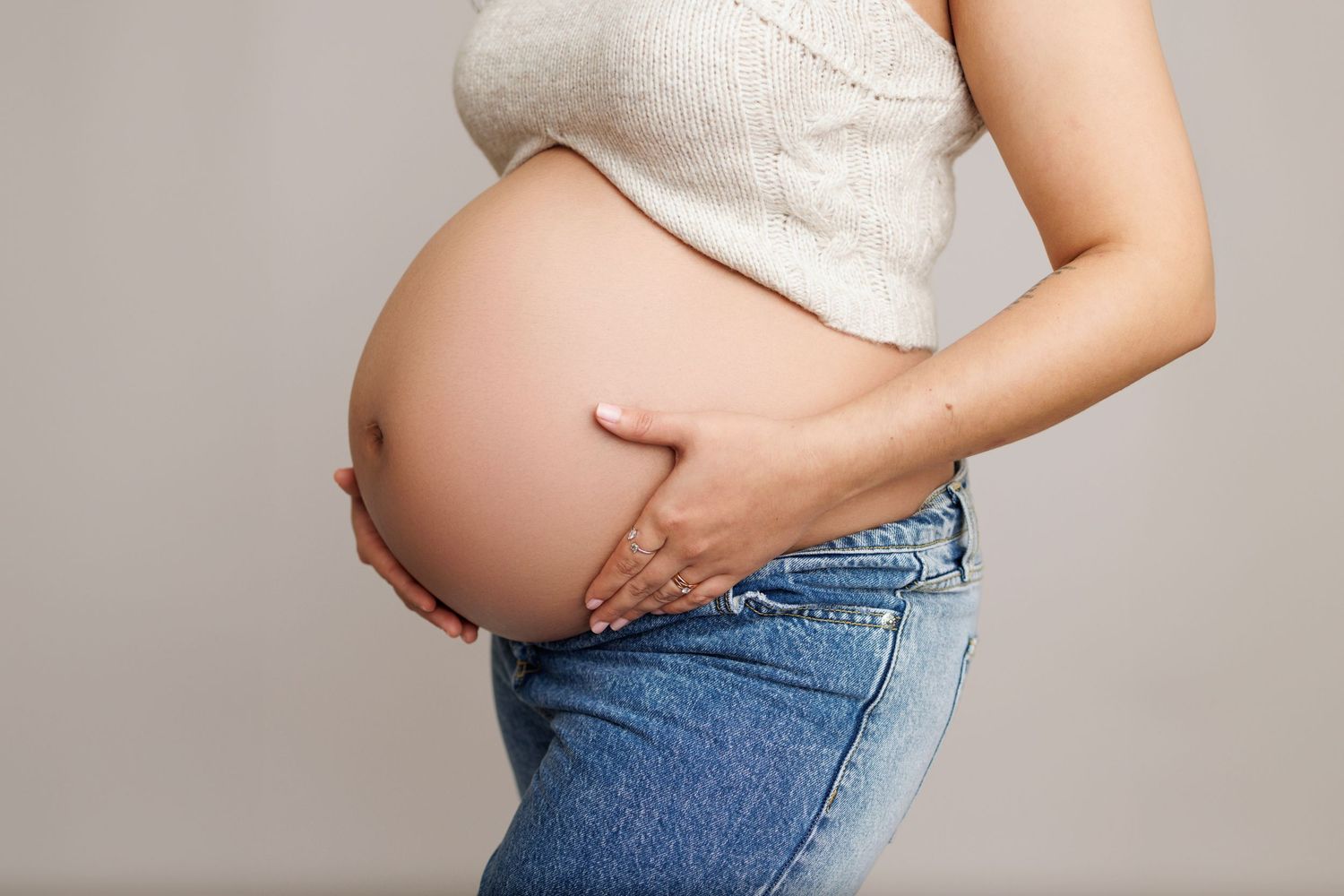 Pregnant belly being cradled by hands while wearing jeans and white top against gray background.
