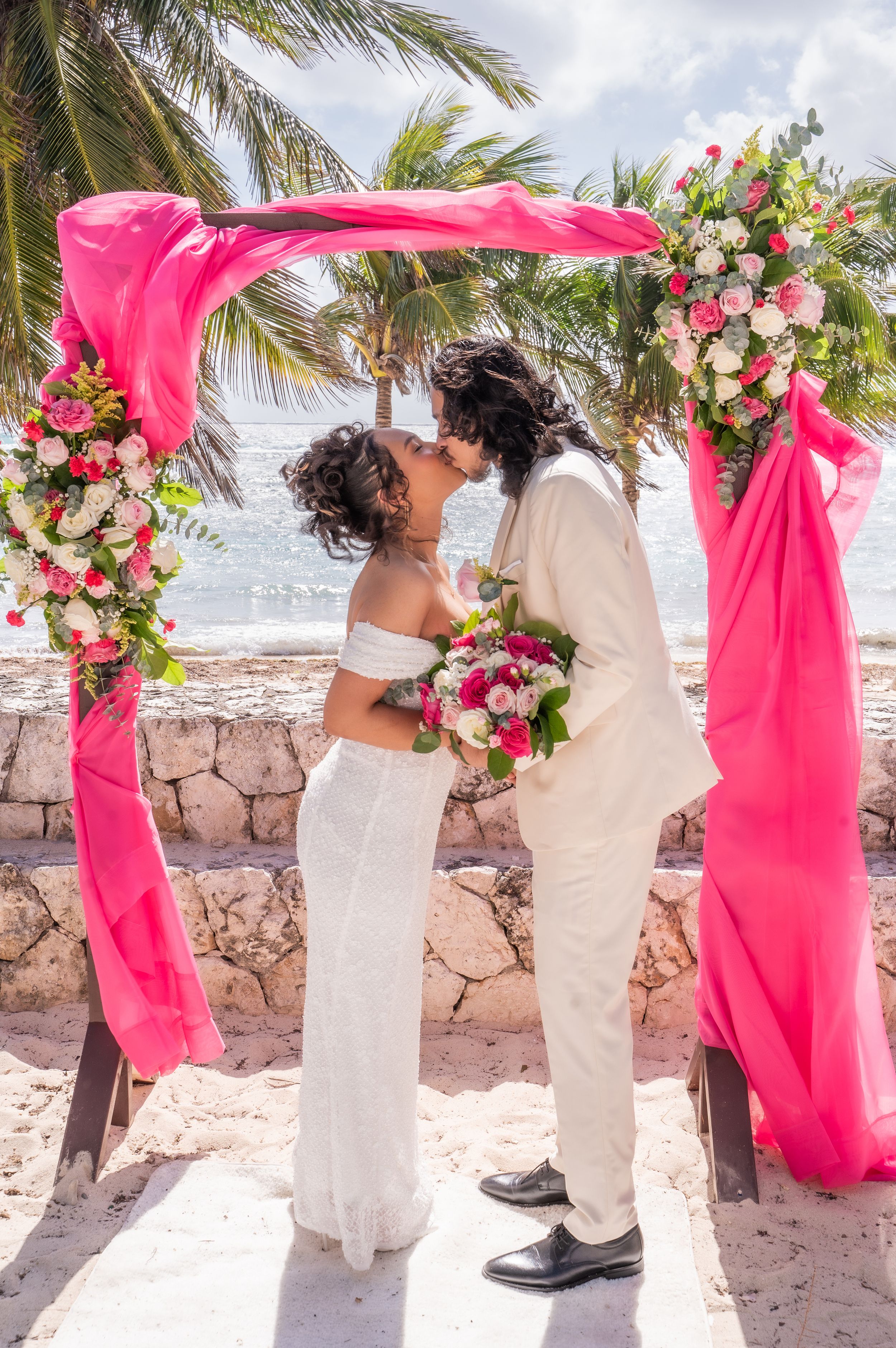 Couple sharing a kiss under a pink floral arch on a beach wedding ceremony with palm trees.